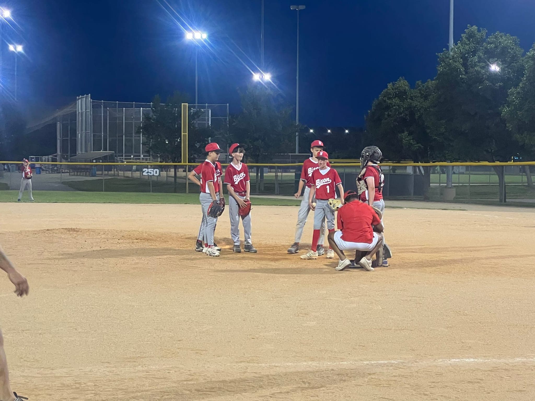 A youth baseball team meeting on the mound under the lights